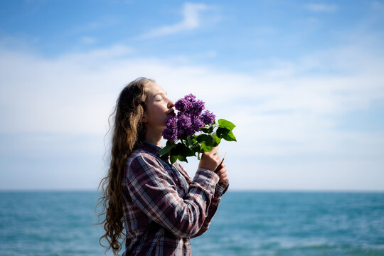 Young woman smelling blooming lilac flowers on a sunny seaside, enjoying nature and a peaceful outdoor moment. Coastal lifestyle, mindfulness and simple pleasures concept.