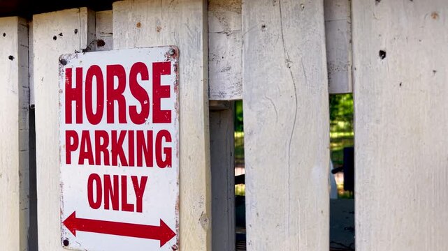 Weathered Horse Parking Only sign with red lettering and double arrow hangs on rustic white wooden fence. Equestrian warning marker creates humorous rural detail at stable, ranch or riding area