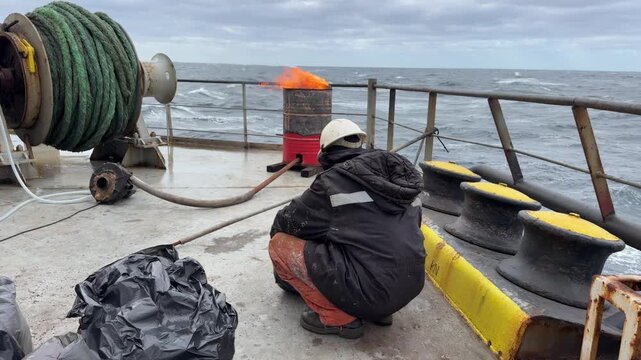 Cargo ship stern view with a worker burning plastic and trash in a special barrel, moving across the wavy sea to reduce environmental footprint.