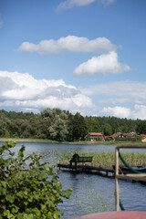 Fototapeta premium Solitary person sitting on a bench at the end of a narrow wooden pier, looking across a peaceful lake toward a rural village under white summer clouds.