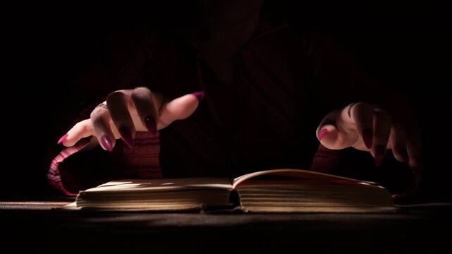 Witch hands casting spells over magic book in dark room close up