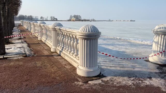 Classic white stone balustrade with red and white warning tape along a frozen snow-covered waterfront park promenade during a sunny winter day.
