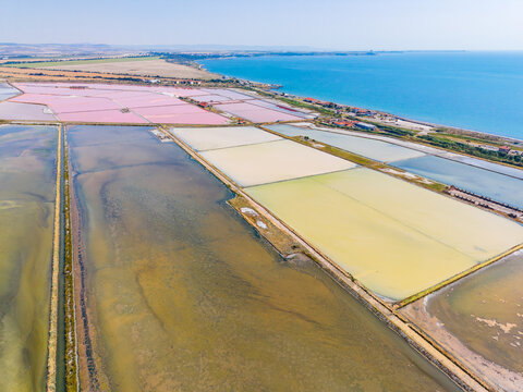 Salt pans stretch across a coastal area with colors changing from pink to yellow under a clear sky during daylight