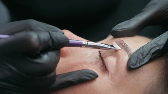 Permanent makeup artist applies white mapping paste to outline eyebrow shape before a cosmetic tattoo procedure in a professional beauty studio.