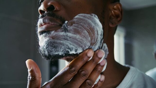Applying man in white tee rubbing shaving foam across face preparing to shave at bathroom sink
