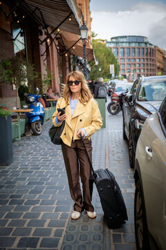 Stylish mature woman with suitcase using smartphone on city street