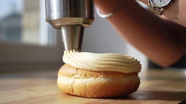 Pressing cook's hand piping decorative cream onto bun at counter, with metal nozzle and wristwatch