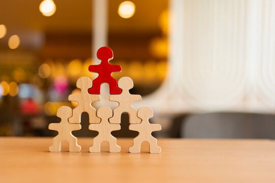 Leadership and business management concept with red wooden figure on top of human pyramid structure on table for career promotion, corporate hierarchy, executive success and teamwork.