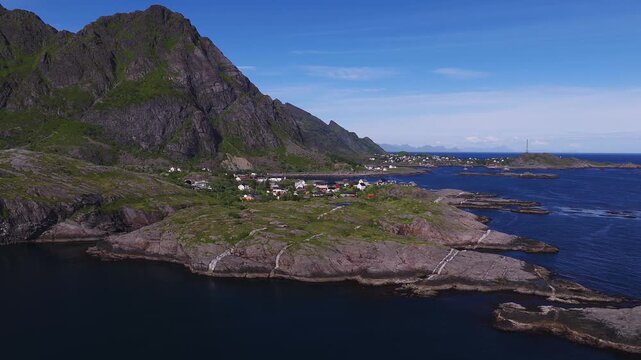 Aerial view reveals a rocky coastline, tranquil fjord waters, and a village nestled beneath rugged mountains in Lofoten, Norway during daylight.