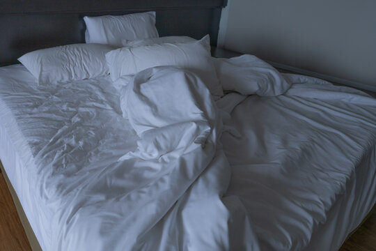 Unmade hotel bed with rumpled white sheets, pillows and duvet in dim guest room after sleep. Everyday travel accommodation, morning mess, comfort, rest and realistic hospitality lifestyle. Lodging.