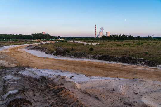 Jaworzno, Poland - 23 August 2026: View of the Jaworzno Power Plant with its cooling towers and chimney rising above a sandy, unpaved road and open field at dusk.
