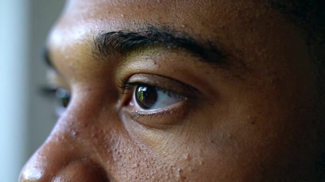 Black man opening left eyelid near window, reacting to bright light, revealing brown iris and pores
