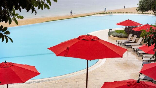 Framing red umbrellas with blue pool on resort deck, two walkers entering left moving along beach