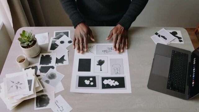 Lifting man wearing dark long-sleeve sweater arranging prints at home studio table, centering sheet