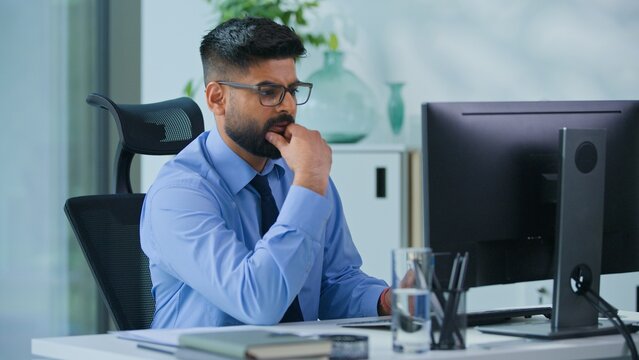 Pensive Indian business man Arabian businessman working with computer looking at screen monitor hesitate Hindu male professional agent thinking pondering work online think solution problem in office
