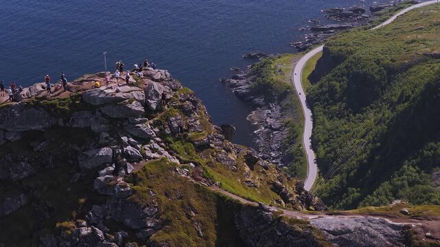 Aerial drone shot of people standing atop the rocky summit of Reinebringen overlooking the coastline road and ocean in Lofoten, Norway.