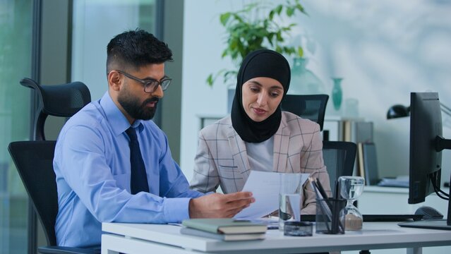 Two concentrated Indian business colleagues discussing papers graphs financial data results talking working computer in office. Focused Arabian man businessman talk team work with woman businesswoman