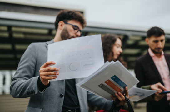 Three business professionals examine printed reports and charts while standing outdoors. Colleagues review documents and data together during an informal urban meeting.