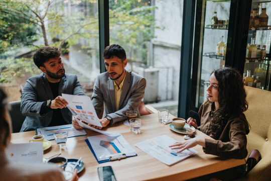 A small group of business colleagues reviews printed reports and discusses strategy over coffee. The team collaborates on documents and charts during a casual meeting at a cafe.