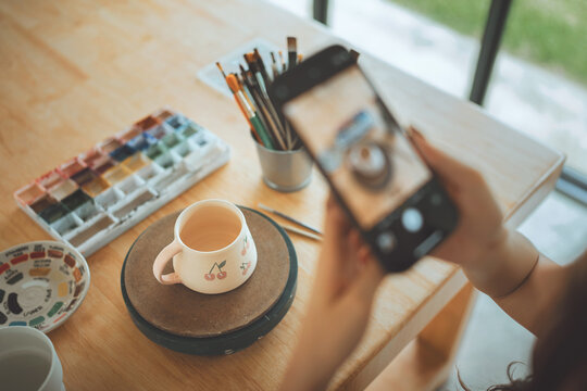 Woman photographing handmade ceramic mug with smartphone in pottery studio, creative craft and artistic process