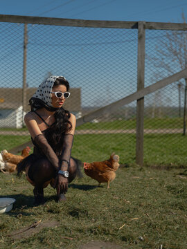 Glamorous Asian woman crouching near chickens on farm yard