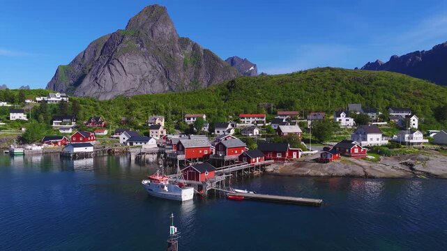 Aerial view of traditional red rorbu cabins and fishing boats along the coast of Reine village in Lofoten, Norway, backed by steep mountain peaks.