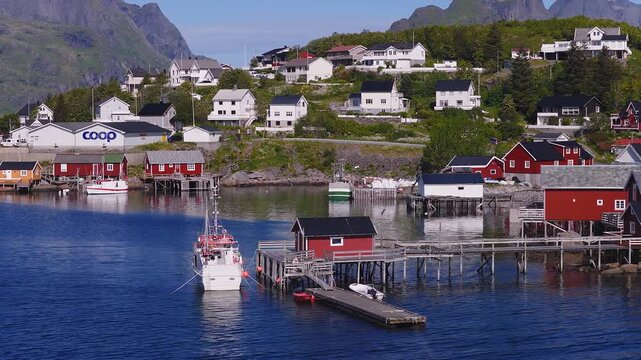 Aerial view of a coastal fishing village in Reine, Lofoten, Norway, showing traditional waterfront houses, wooden piers, moored boats, and mountains.