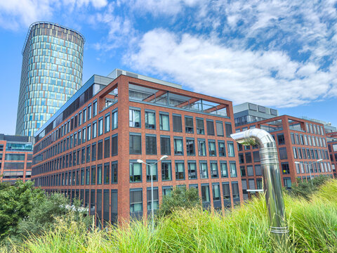 Modern buildings stand in a city with a shiny pipe in the foreground under a blue sky with clouds