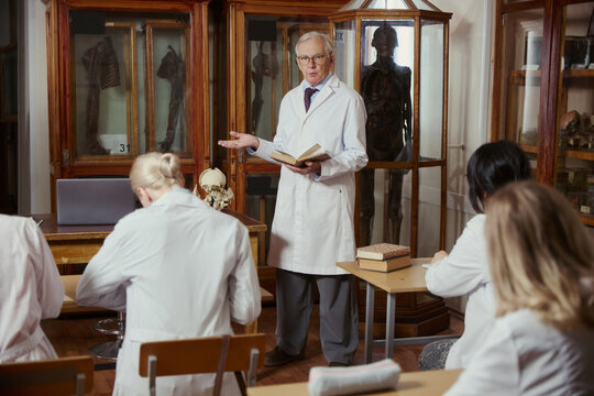 Senior Caucasian male professor holding book and lecturing medical students in anatomy museum. Image suitable for university education, medical training, and academic marketing