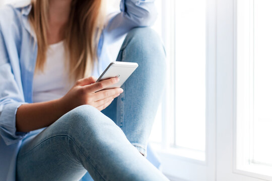 Woman using smartphone at home, sitting by window. Girl browsing, shopping online. Distance learning, remote education, connection, communication. Doomscrolling, mobile addiction. Close up of hands