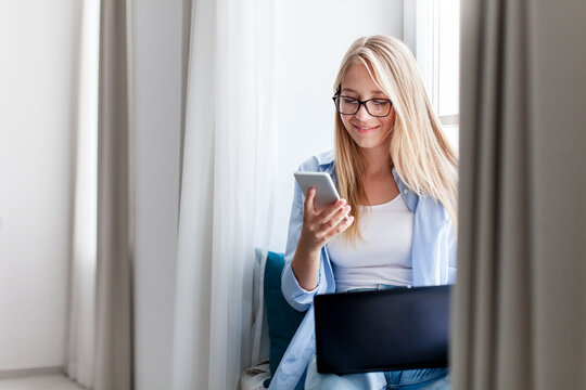 Young woman using laptop, smartphone, working from home office. Freelancer workplace by window. Technology lifestyle, teleworking. Online education, distance learning for student