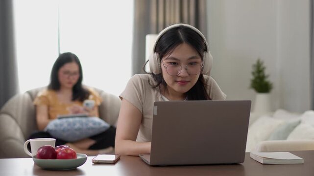A young woman works on a laptop at home while another woman sits in the background. The footage conveys concentration, remote work, digital productivity, and a contemporary home lifestyle.
