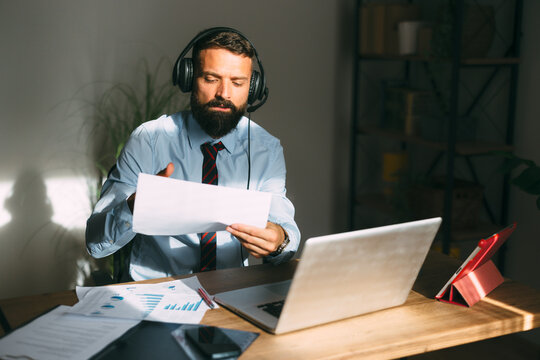 Man with a beard wearing a headset with a microphone, holding documents and looking at a laptop screen during an online video conference from his home office, embracing the new normal of remote work