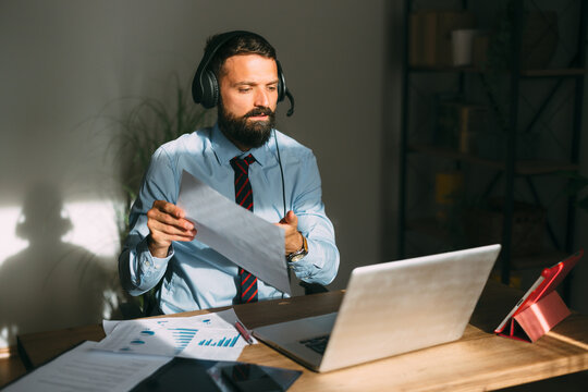 Businessman wearing a headset with microphone participating in a video call, reviewing documents and working on a laptop from his home office, highlighting remote work and online communication
