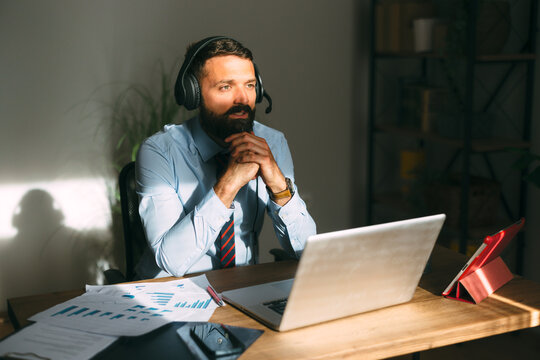 Bearded businessman wearing a shirt and tie conducting a video call with a headset and laptop at a desk, engaging in online consulting or virtual meeting from a home office