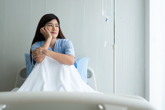 Young woman hospital patient with severe headache lies in a medical bed while wearing a face mask and holding her head, illustrating illness, pain, recovery, healthcare treatment and clinical.