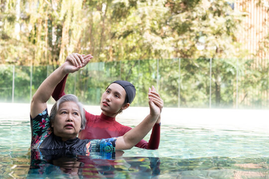 Senior woman receiving aquatic therapy guidance from a young female instructor in a swimming pool, lifting both arms through controlled movements that support healthy aging and balanced wellness.