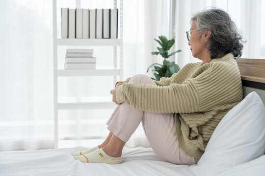 Senior woman sitting on a bed with a concerned expression in a bright bedroom, representing emotional wellbeing, aging, healthcare, and retirement lifestyle themes for wellness and counseling cam