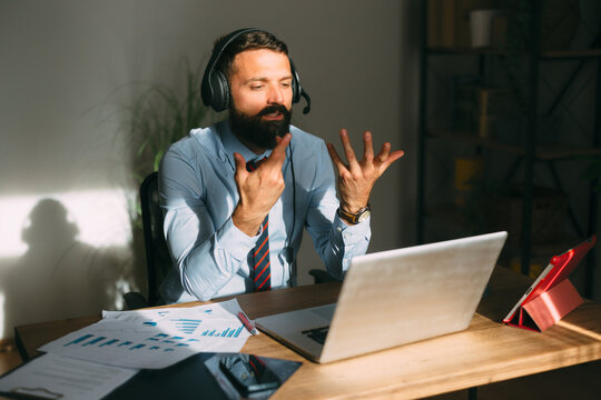 Bearded man wearing a headset and formal shirt engaged in an online video conference, using hand gestures while discussing business, working remotely from his sunlit home office desk