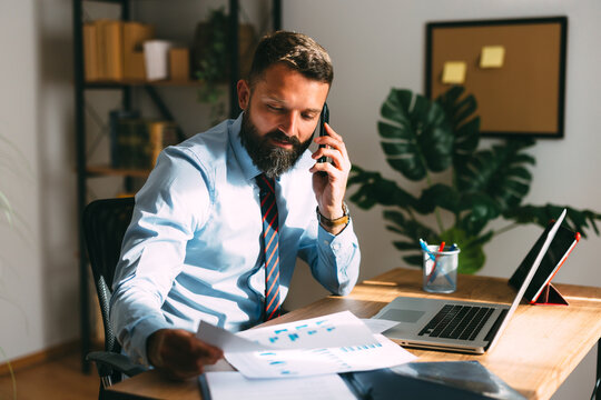 Male professional working from a home office, talking on a smartphone while reviewing business documents and financial reports with a laptop and digital tablet on his desk