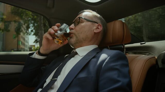 Middle aged Caucasian businessman sitting in back seat of luxury car, adjusting collar and drinking whiskey. Footage is ideal for corporate videos or luxury brand advertisements