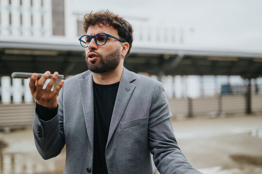 A young businessman speaks into his smartphone holding it at arm&rsquo;s length. He uses voice messaging or makes a call while standing outdoors in a city environment.