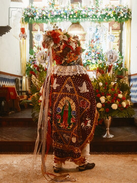 Angahuan, Michoacan, Mexico - 24 July 2026: View of a Moros dancer in traditional costume with floral headdress and Virgin of Guadalupe cape standing before a decorated church altar.