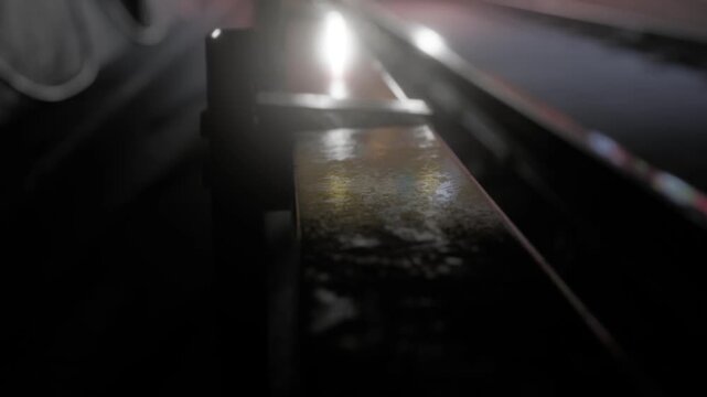 A close up view of a rusty metal rail shows light reflections. The setting is dark, highlighting the textures of the metal. This could be near a train platform or factory area at night.