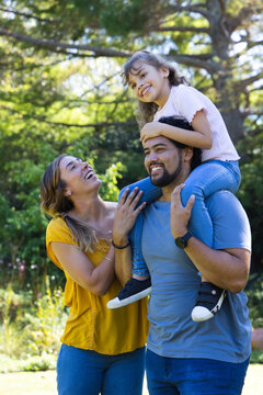 Family posing in park under trees father carrying daughter on shoulders mom laughing sneakers watch