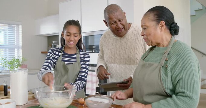 African family using tablet guiding girl cracking eggs whisking batter with sieve at kitchen island