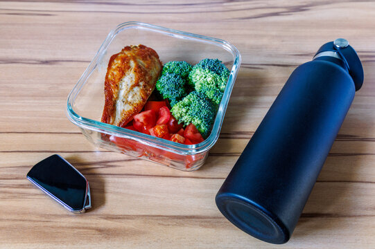 Glass container with roasted chicken breast, broccoli and tomatoes near black water bottle and car key on wooden table, Healthy lunch prep and daily commuting concept