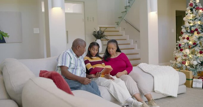 African American family on sofa teenager reading album after gift near tree, dad pointing, laughing