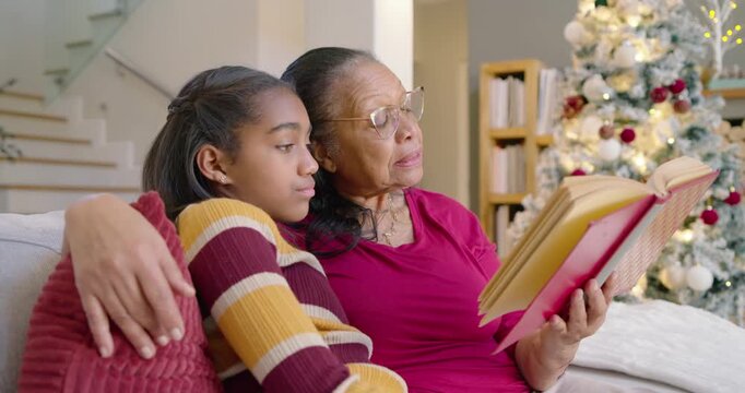 African American senior woman opening red book while youth girl leaning on sofa, bonding