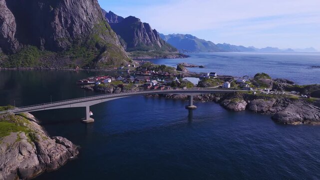 Aerial drone view across the concrete bridge connecting rocky islands and traditional village houses in Lofoten, Norway on a clear sunny day.